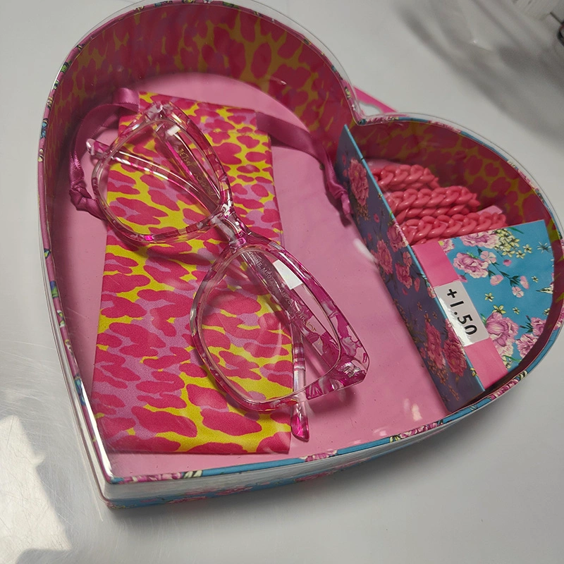 Heart-shaped gift box containing a pair of transparent pink reading glasses, a matching pink and yellow patterned cleaning cloth, and a floral-patterned section with pink accessories, labeled +1.50.