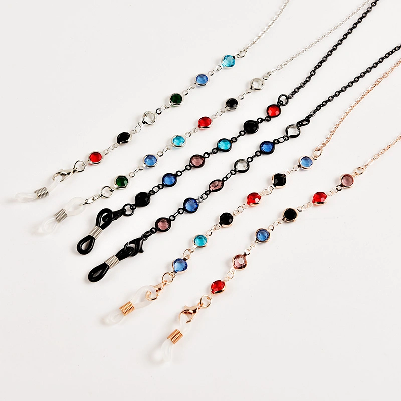 A top-down view of five decorative glasses chains spread diagonally on a white background. The chains come in silver, black, and rose gold finishes, each one adorned with small, colorful, bezel-set rhinestones in various colors like blue, red, green, and pink. Each chain has an adjustable rubber loop at the end to attach to eyewear.