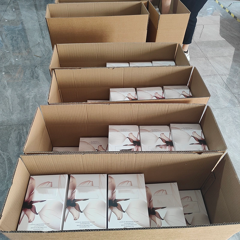 Open cardboard boxes neatly arranged in rows on a tiled floor, each box filled with multiple pairs of sunglasses in white packaging adorned with a soft floral magnolia-petal design and a rectangular transparent window revealing the sunglasses inside.