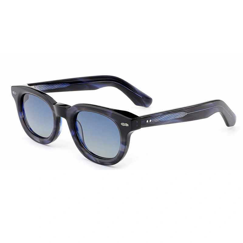 A studio shot of a pair of thick-frame sunglasses with a dark blue and black marbled pattern, shown from a three-quarter angle on a white background. The sunglasses feature round, blue gradient lenses and thick temples with visible silver hinge details.